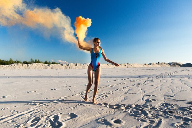 Chica en traje de baño azul baila con humo naranja en la playa blanca