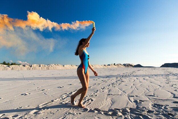 Chica en traje de baño azul baila con humo naranja en la playa blanca