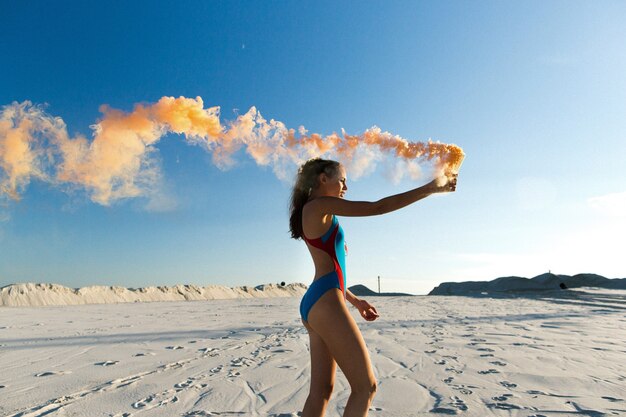 Chica en traje de baño azul baila con humo naranja en la playa blanca