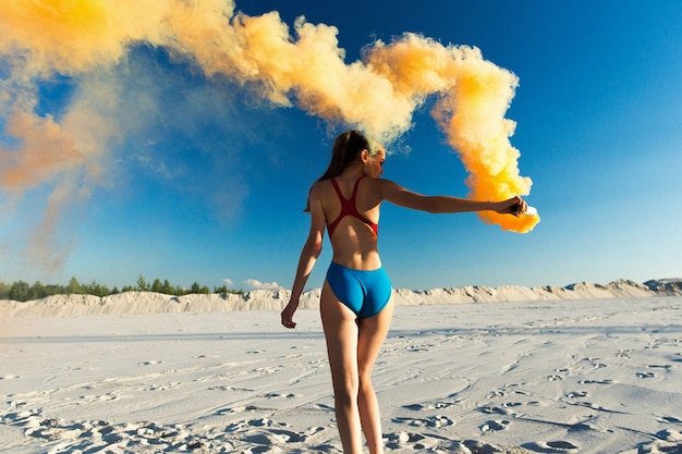 Chica en traje de baño azul baila con humo naranja en la playa blanca