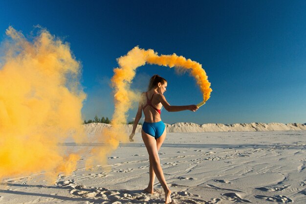 Chica en traje de baño azul baila con humo naranja en la playa blanca