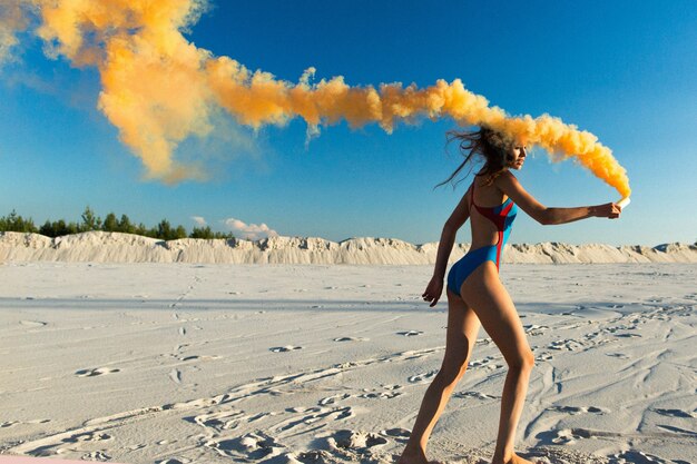 Chica en traje de baño azul baila con humo naranja en la playa blanca