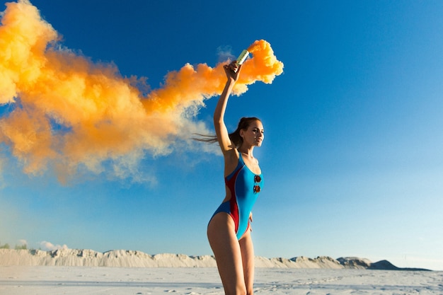 Chica en traje de baño azul baila con humo naranja en la playa blanca