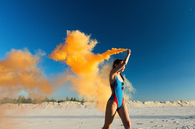 Foto gratuita chica en traje de baño azul baila con humo naranja en la playa blanca