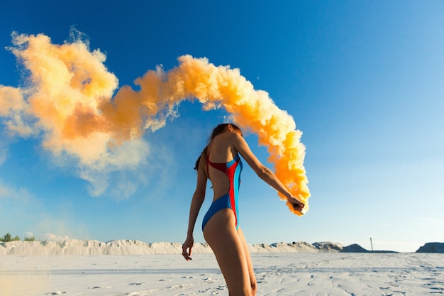 Foto gratuita chica en traje de baño azul baila con humo naranja en la playa blanca