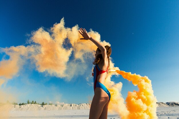 Chica en traje de baño azul baila con humo naranja en la playa blanca