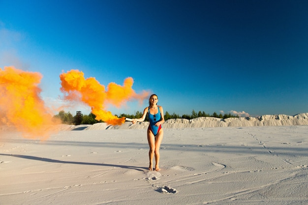 Chica en traje de baño azul baila con humo naranja en la playa blanca