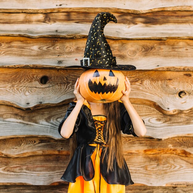 Chica en traje de bruja que cierra la cara por calabaza con sombrero