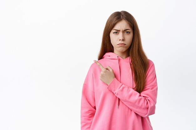 Chica triste y decepcionada deja de lado, frunce el ceño y se ve celosa, se queja de algo injusto, de pie contra la pared blanca