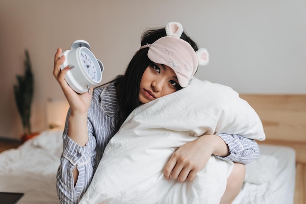 Chica triste en pijama y antifaz para dormir está abrazando una almohada blanca y sosteniendo un despertador