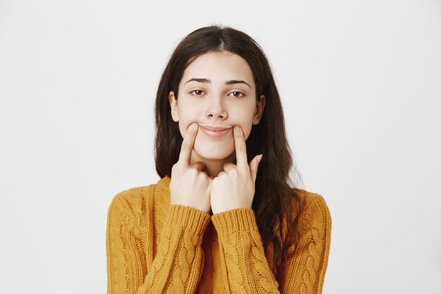 Chica triste tratando de tirar de una sonrisa feliz, empujando las esquinas de los labios hacia arriba con cara reacia