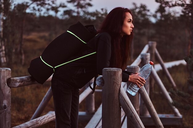 Chica turista con una capucha negra y jeans sostiene un agua y se apoya en una cerca de madera en un hermoso prado de otoño al atardecer.