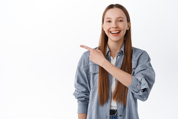 Chica universitaria alegre con cabello largo apuntando a un lado sonriendo y mostrando el logo de esta manera gesto recomendando hacer clic en la pancarta riendo feliz de pie sobre fondo blanco