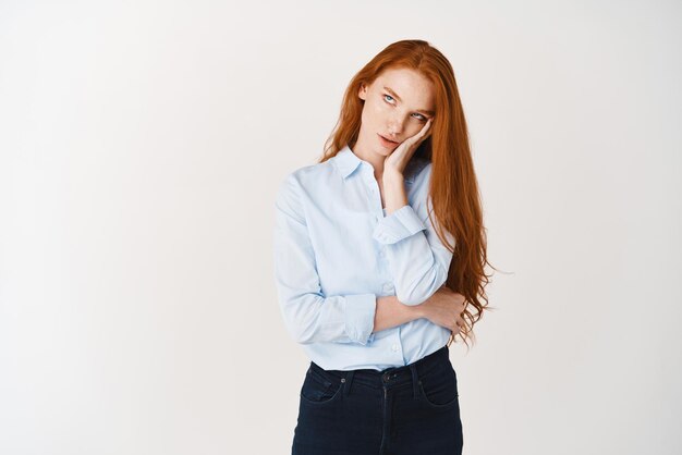 Chica universitaria enfadada con el pelo largo y rojo de pie cabreada, pone los ojos en blanco y expresa aburrimiento o arrogancia de fondo blanco