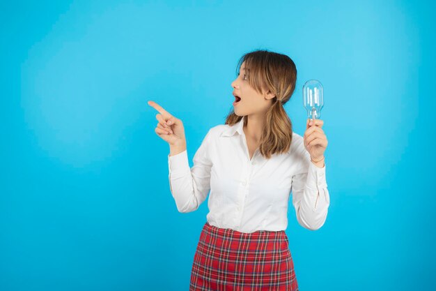 Chica universitaria sorprendida sosteniendo una bombilla de idea y señalando con el dedo hacia el lado derecho. foto de alta calidad