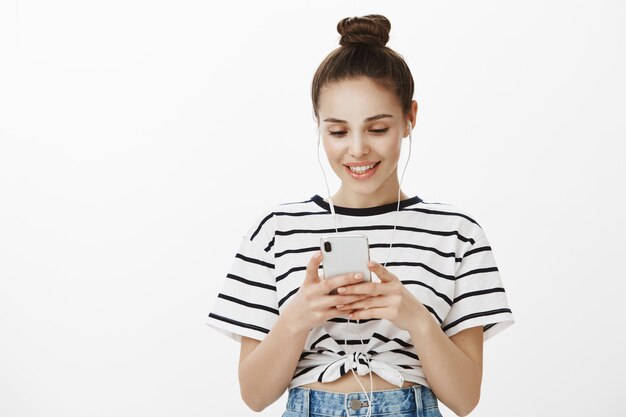 Chica urbana con estilo en auriculares, sonriendo y mirando la pantalla del teléfono inteligente complacido