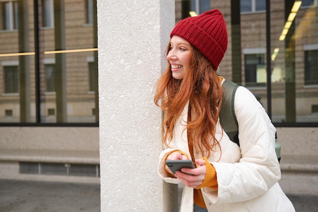 Foto gratuita chica urbana con estilo usando la aplicación de teléfono móvil de pie en la ciudad esperando un taxi mirando la aplicación del teléfono inteligente