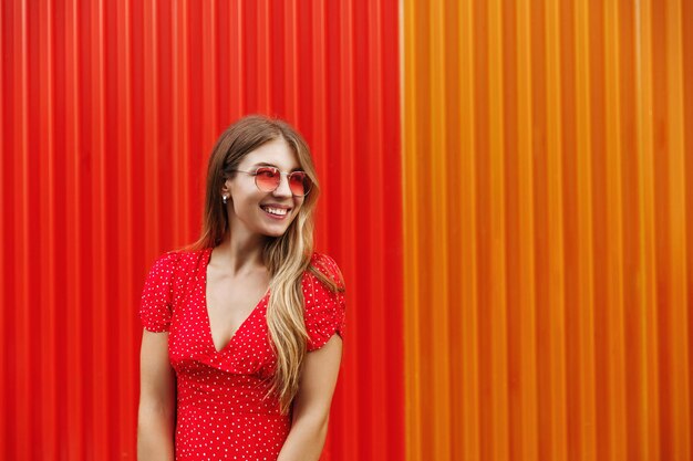 Chica urbana feliz con gafas de sol y vestido de pie cerca de la colorida pared roja mirando a la derecha con una sonrisa alegre explorar la ciudad en vacaciones de verano