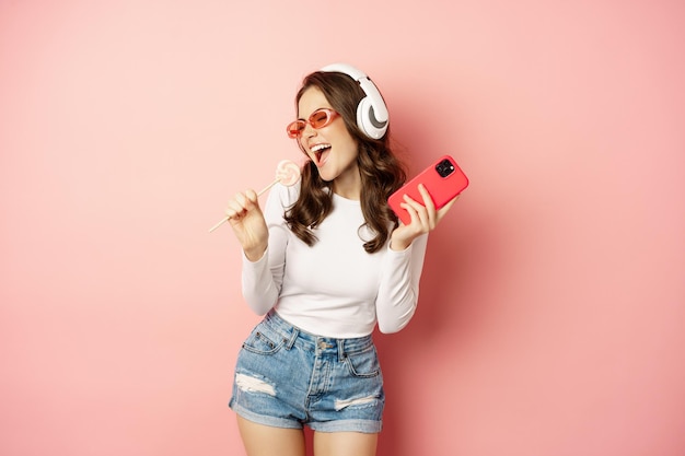 Chica de verano riendo comiendo piruleta y escuchando música en auriculares bailando con el teléfono inteligente otra vez...