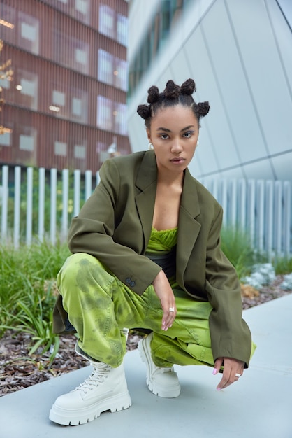 Chica vestida con chaqueta formal verde, pantalones sueltos y botas blancas plantea contra edificios urbanos al aire libre