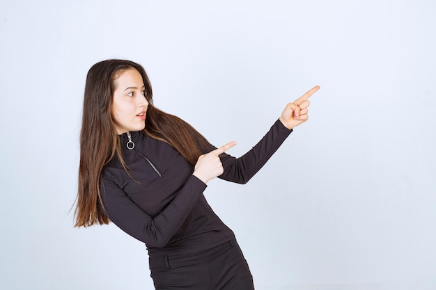 Chica vestida de negro apuntando a algo a la derecha.