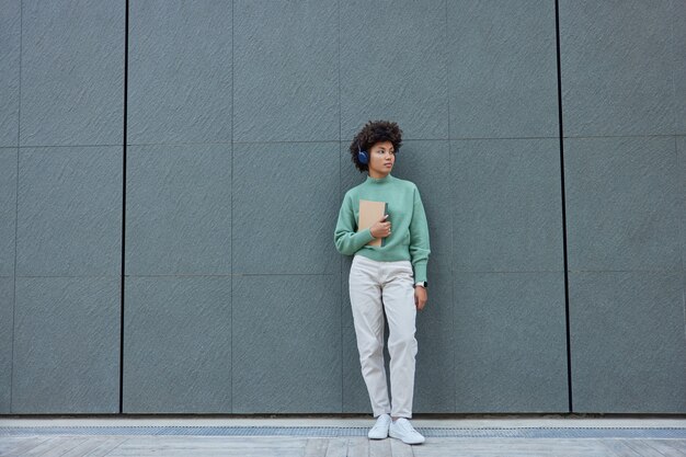 Chica vestida con ropa de moda mira hacia otro lado escucha música en auriculares sostiene poses de diario contra la pared gris