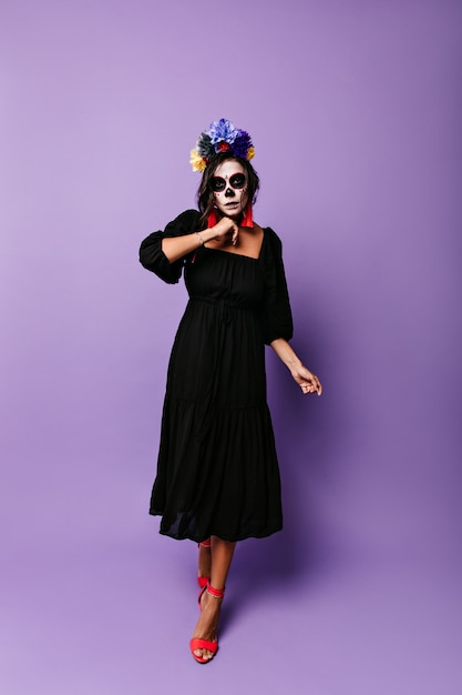 Chica en vestido midi negro camina contra la pared púrpura. Modelo con máscara de calavera en poses de cara para foto de Halloween.