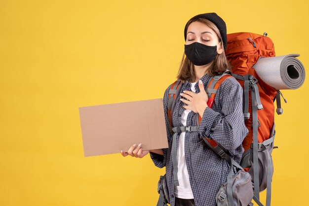 Chica viajera con máscara negra y mochila poniendo la mano sobre su pecho sosteniendo cartón