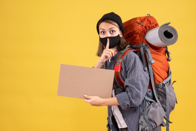 Chica viajera con máscara negra y mochila sosteniendo cartón haciendo letrero de silencio