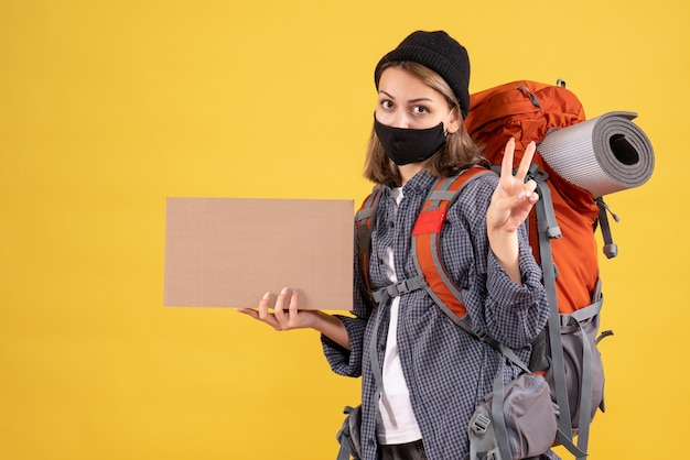 Chica viajera con máscara negra y mochila sosteniendo cartón haciendo el signo de la victoria