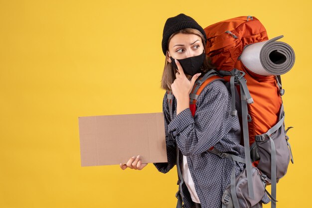 Chica viajera con máscara negra y mochila sosteniendo cartón mirando a la derecha