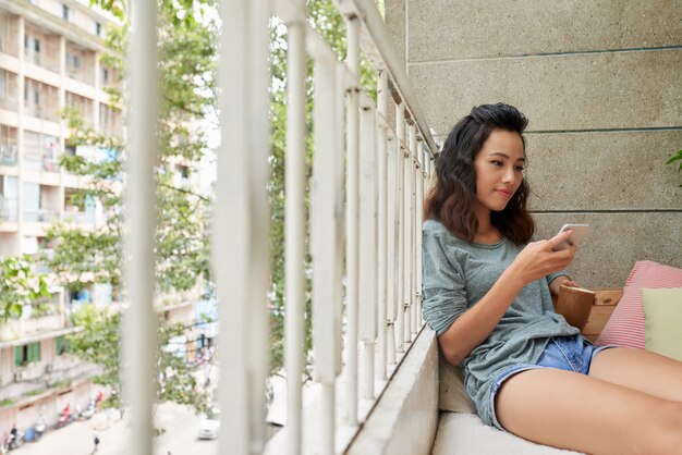 Chica vietnamita atractiva enviando mensajes de texto y tomando té en el balcón