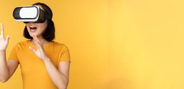 Chica en VR Hermosa joven asiática usando gafas de realidad virtual y jugando chateando virtualmente de pie sobre fondo amarillo