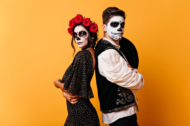 Chica zombie bien formada en vestido negro posando con su novio. Foto interior de una pareja guapa con maquillaje de calavera.