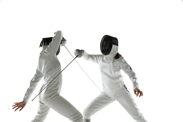 Chicas adolescentes en trajes de esgrima con espadas en manos aisladas sobre fondo blanco de estudio. Modelos femeninos jóvenes practicando y entrenando en movimiento, acción. Copyspace. Deporte, juventud, estilo de vida saludable.