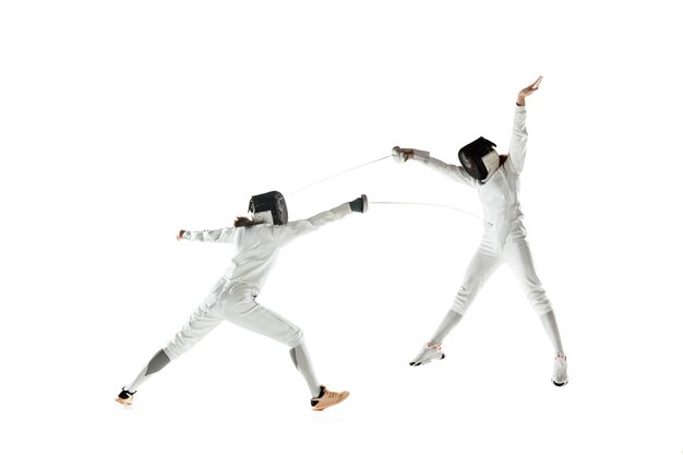 Chicas adolescentes en trajes de esgrima con espadas en manos aisladas sobre fondo blanco de estudio. Modelos femeninos jóvenes practicando y entrenando en movimiento, acción. Copyspace. Deporte, juventud, estilo de vida saludable.