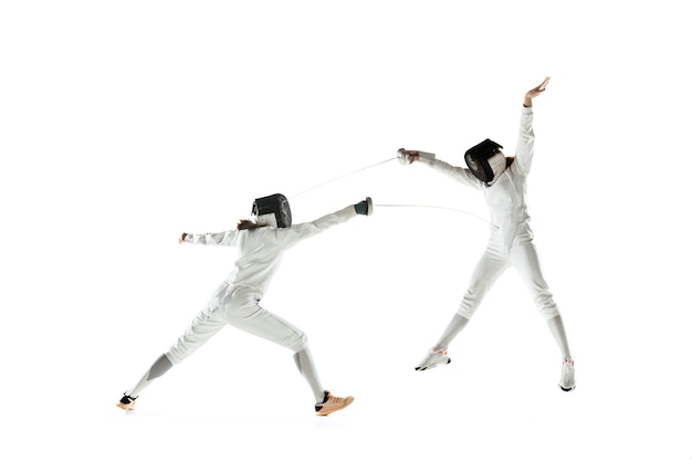 Chicas adolescentes en trajes de esgrima con espadas en manos aisladas sobre fondo blanco de estudio. Modelos femeninos jóvenes practicando y entrenando en movimiento, acción. Copyspace. Deporte, juventud, estilo de vida saludable.