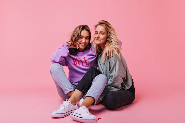 Chicas adorables sentadas con las piernas cruzadas y abrazándose. Retrato interior de hermosas amigas posando en el suelo.