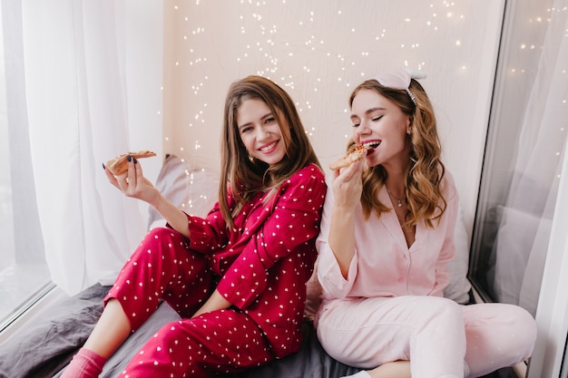 Chicas agraciadas sentadas en la cama y comiendo pizza. Señoras bastante caucásicas que pasan la mañana del fin de semana en el dormitorio con comida rápida.