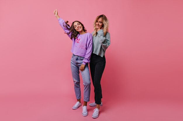 Chicas alegres delgadas riendo durante la sesión de retratos de estudio. Retrato interior de amigas emocionales aisladas en rosa.