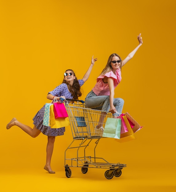 Chicas alegres y felices con muchas bolsas de compras sentadas en un carrito de compras aisladas sobre el fondo amarillo del estudio
