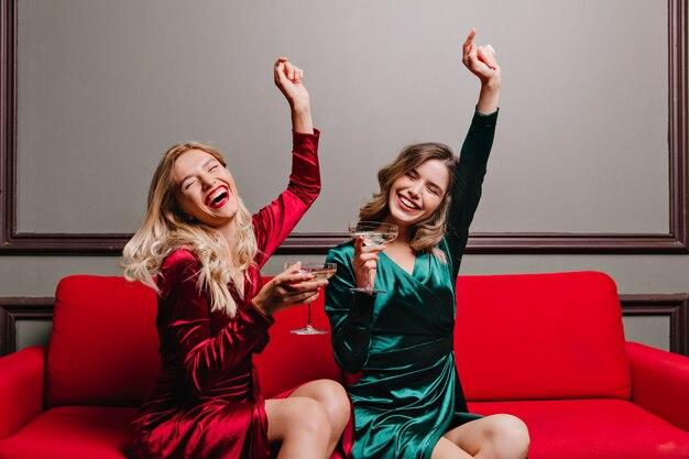 Chicas alegres vestidas de terciopelo bebiendo vino. Retrato interior de glamorosas damas sentadas en el sofá con copas de champán.