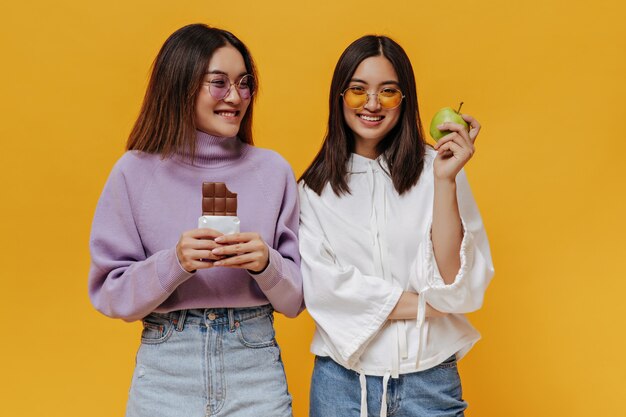 Chicas atractivas en gafas de sol de colores posan en la pared naranja aislada. Chica en suéter morado tiene barra de chocolate con leche