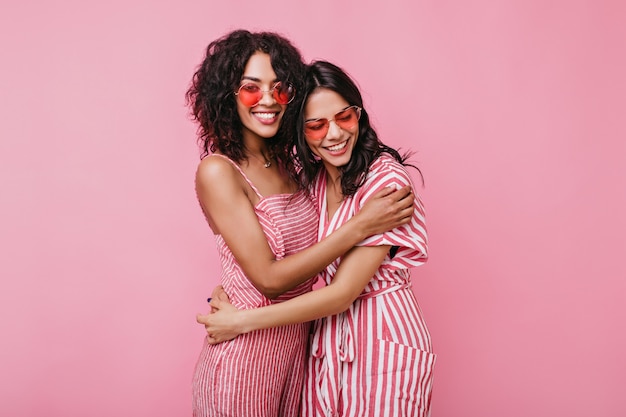 Las chicas bronceadas con sinceras sonrisas blancas se regocijan en las sesiones de fotos. Novias en monos de verano posando.