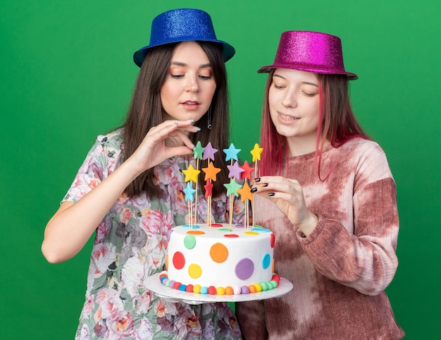 Chicas complacidas con gorro de fiesta sosteniendo y mirando pastel aislado en la pared verde