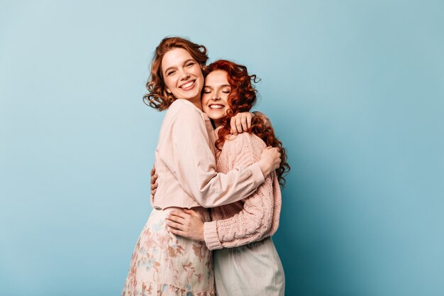 Chicas debonair abrazando sobre fondo azul. Foto de estudio de amigos atractivos que se ríen de la cámara.