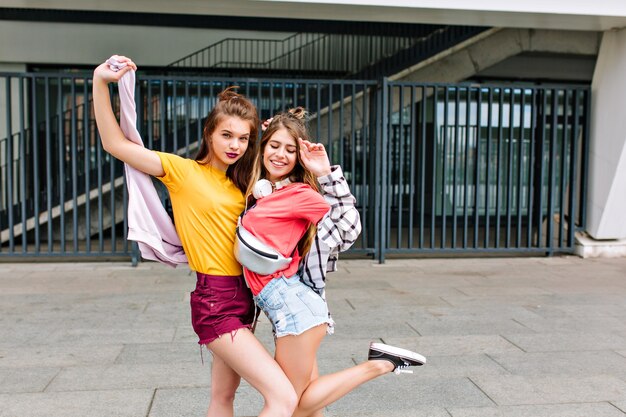 Chicas delgadas en lindos pantalones cortos de mezclilla bailando frente a la valla de hierro y sonriendo disfrutando el fin de semana