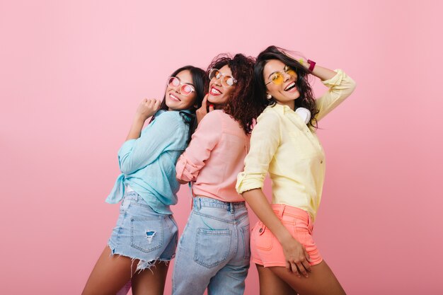 Chicas despreocupadas con coloridas camisetas de algodón posando juntas y sonriendo. Retrato interior de atractivas señoritas que expresan emociones felices.