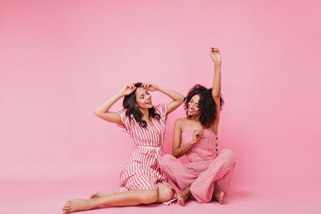 Chicas dulces y encantadoras con cabello negro y rizado, en pijama rosa divirtiéndose y levantando las manos, bailando