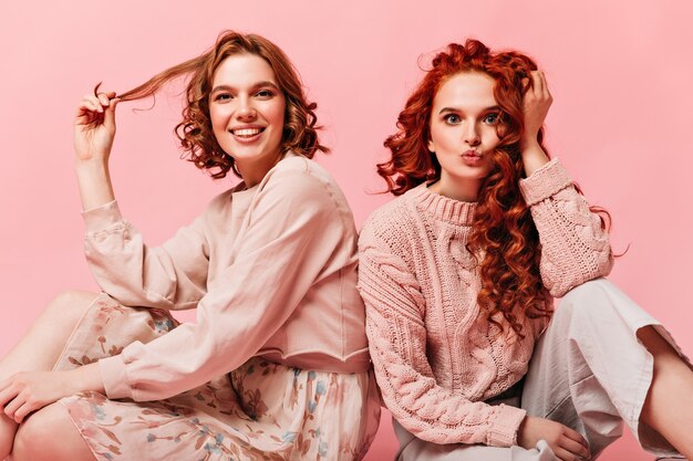 Chicas encantadoras sentadas sobre fondo rosa y tocando el pelo rizado. Vista frontal de amigos posando en el piso.